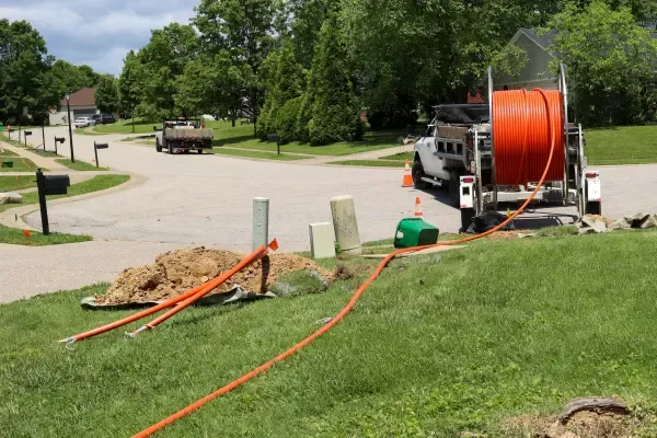 Utility workers installing underground fiber optic cables in a residential neighborhood, with orange conduit tubing laid across lawns and connected to utility boxes.